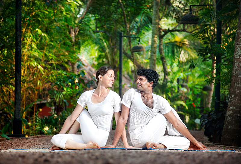 Couple sits in Matsyendrasana yoga pose, looking at each other, both wearing white clothing.