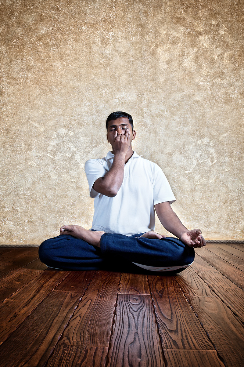 Young man sits cross-legged on wooden floor, eyes closed, using pointer and middle finger to perform Nadi Suddhi pranayama, a yoga breathing technique.