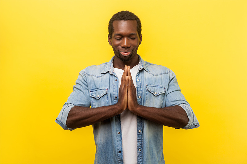 A portrait of a person of color wearing denim jacket, standing with eyes closed in meditative state against a yellow background. 