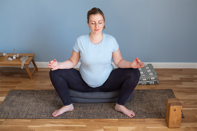 Pregnant Caucasian woman meditating, sitting with legs folded like an upright frog, wearing a blue shirt and tight-fitting denim jeans, with eyes closed and index and thumb resting on her knees.