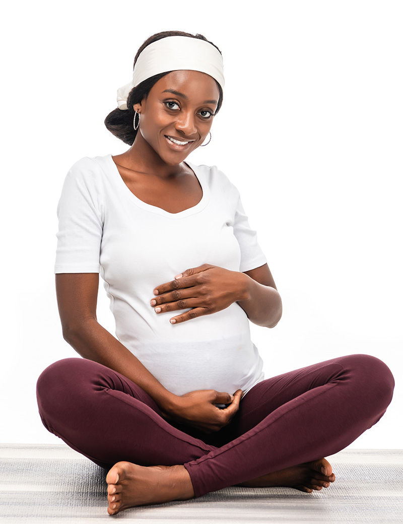 Pregnant woman of color smiling while holding her tummy, wearing a white headband, top, and Bordeaux leggings while sitting cross-legged.